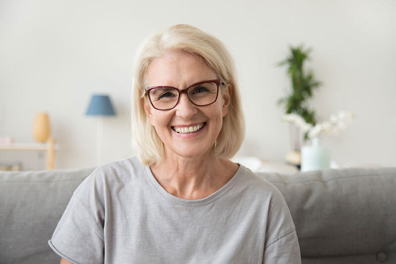 An older woman with glasses smiling on the couch.